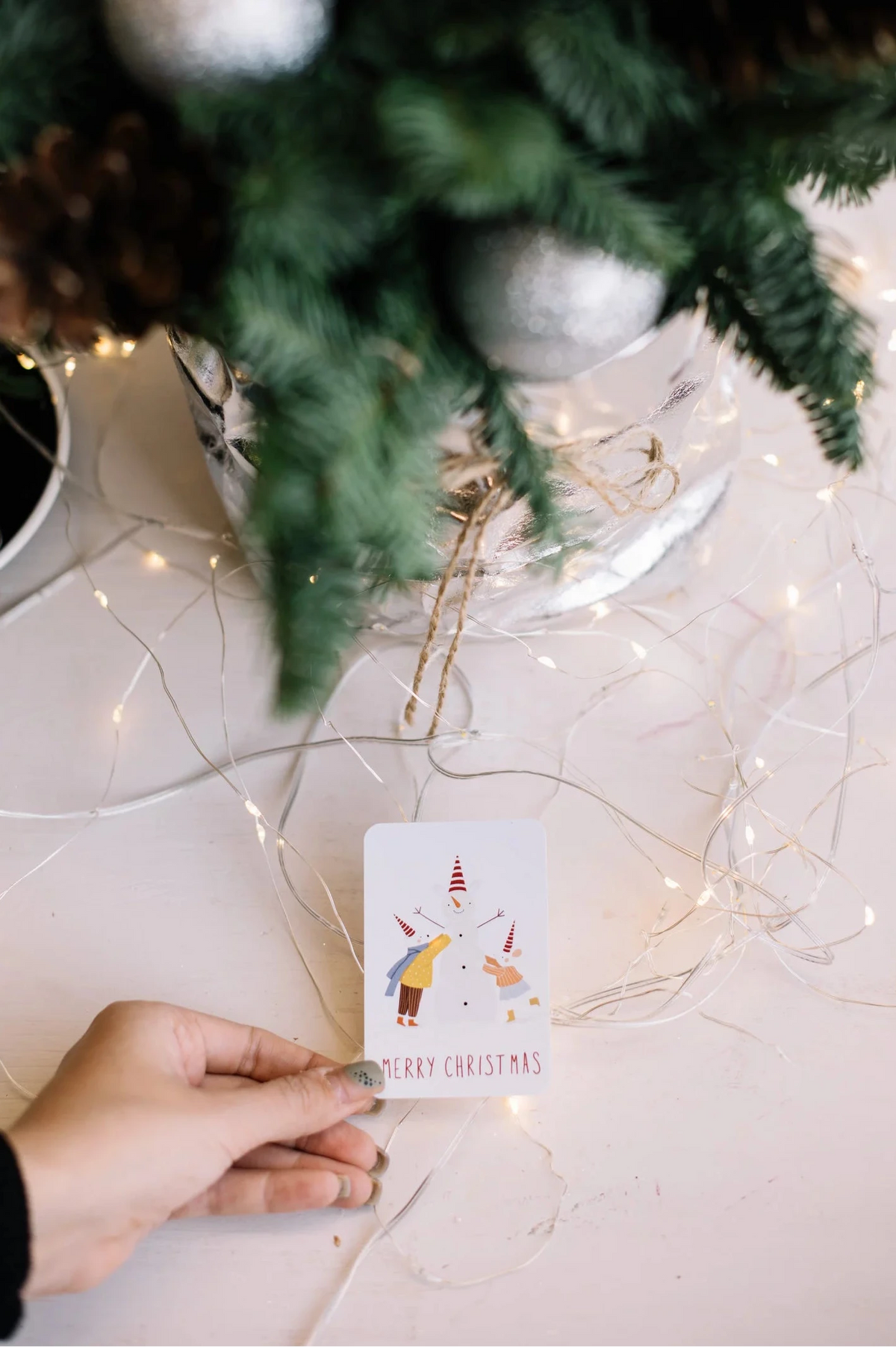 Mini card illustrated with a snowman flanked by two people in winter coats and scarves, with ‘Merry Christmas’ text below, held in hand near a fir branch, string lights, and silver ornaments