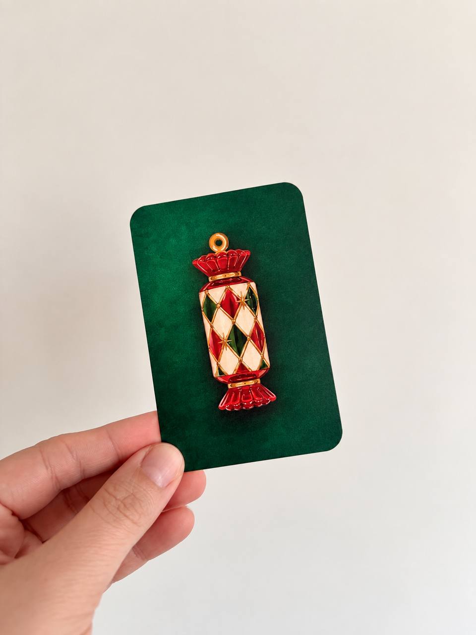 Hand holding a mini green Christmas card with an embossed red, green, and gold jeweled Christmas cracker illustration.