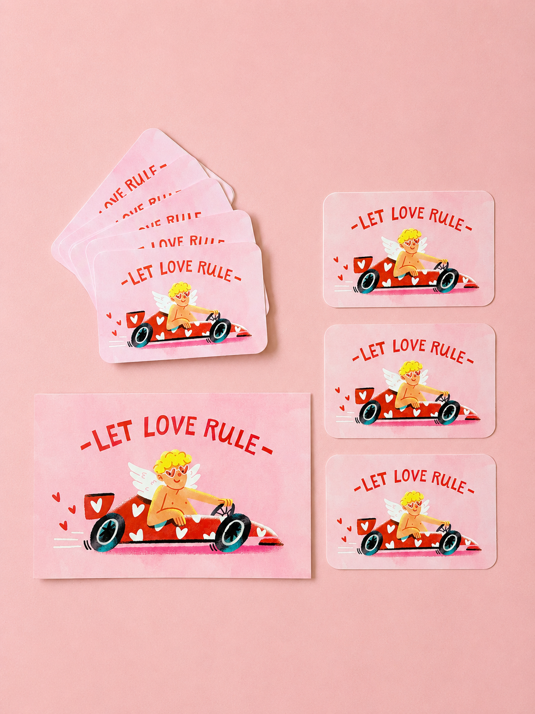 Pink “Let Love Rule” cards featuring a cupid driving a heart-covered red race car, arranged on a soft powder-pink background.