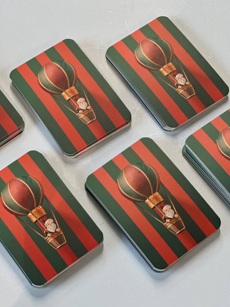 Stacks of mini Christmas greeting cards illustrated with Santa riding a hot air balloon on a red and green striped background, shown in bulk wholesale product photography.