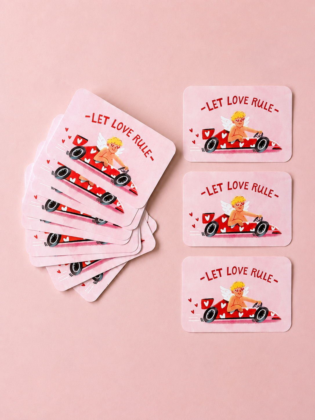 Pink “Let Love Rule” cards featuring a playful cupid driving a red heart-covered race car, arranged on a soft powder-pink background