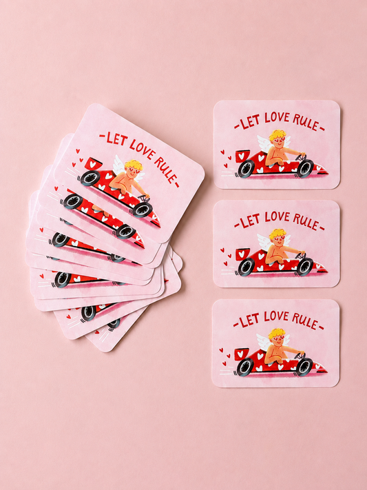 Pink “Let Love Rule” cards featuring a playful cupid driving a red heart-covered race car, arranged on a soft powder-pink background