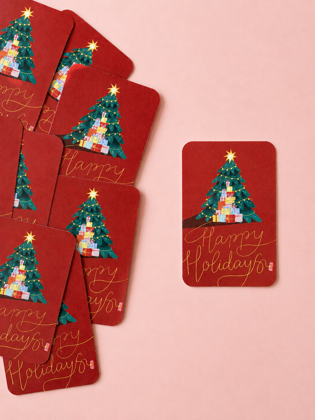 Red “Happy Holidays” mini cards featuring a decorated Christmas tree and colourful gifts, arranged on a soft powder pink background.