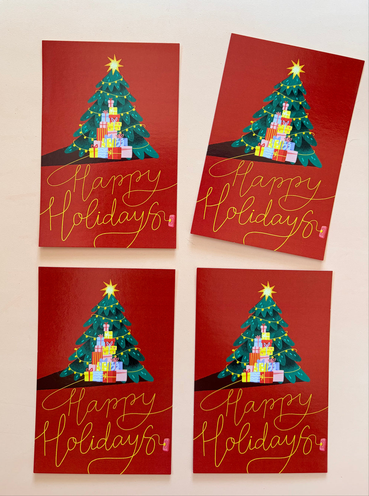 Four red Christmas greeting cards featuring a decorated Christmas tree, colourful presents and gold “Happy Holidays” lettering, arranged on a soft powder-pink background.