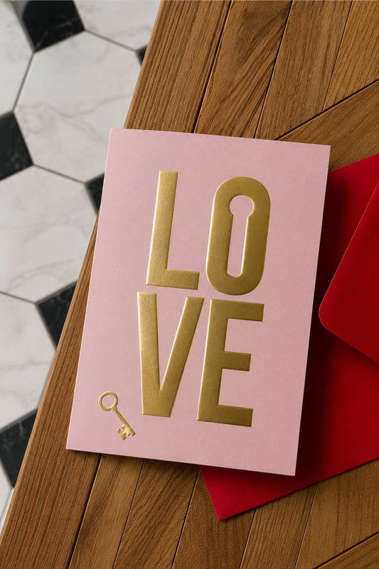 Rose mini greeting card with gold foil embossed "LOVE" lettering and a key icon, paired with a contrasting red envelope on a wooden surface.