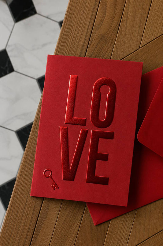 Red mini greeting card with tone-on-tone red foil embossed “LOVE” lettering and a key icon, next to a matching red envelope on a wooden surface.