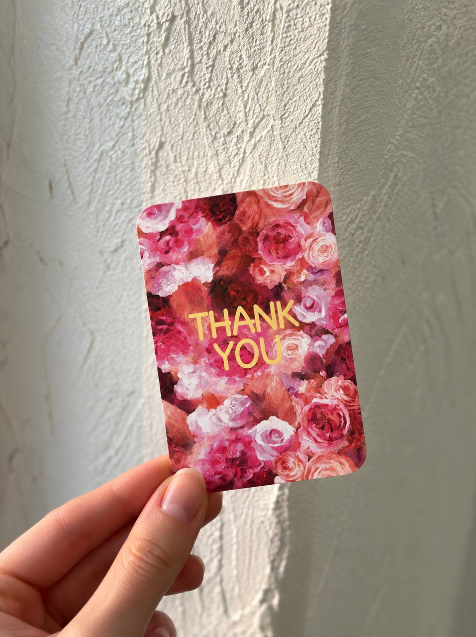 Hand holding a mini greeting card with a dense rose floral illustration and "Thank You" in gold foil, against a textured wall.