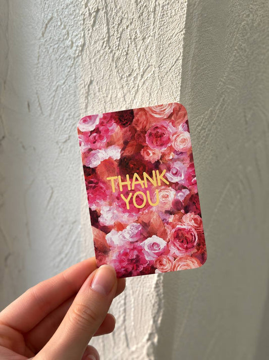 Hand holding a mini greeting card with a dense rose floral illustration and "Thank You" in gold foil, against a textured wall.