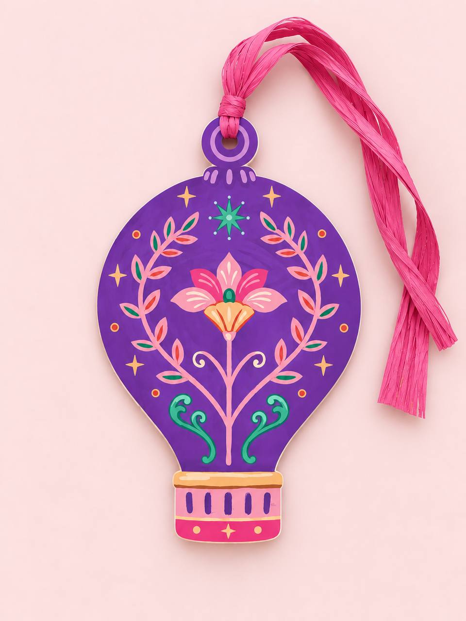 Shaped paper ornament "Talisman Hot Air Balloon" in purple with a pink floral and vine folk-art design, tied with a pink raffia tassel on a pink background.