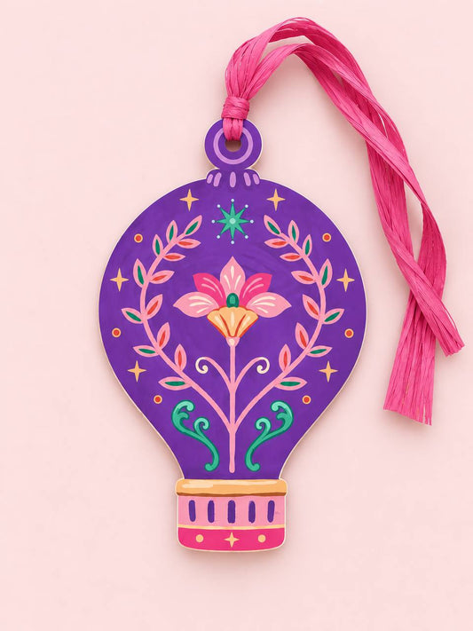 Shaped paper ornament "Talisman Hot Air Balloon" in purple with a pink floral and vine folk-art design, tied with a pink raffia tassel on a pink background.