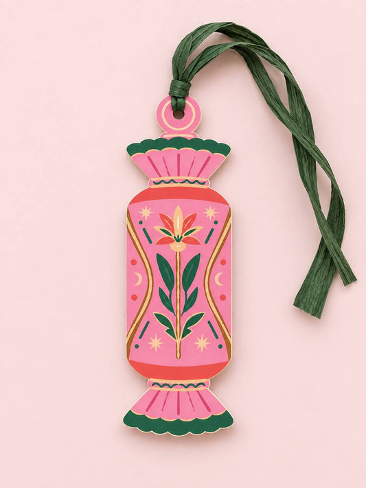 Shaped paper ornament "Talisman Candy" with a pink and orange folk-art floral design, tied with a green raffia tassel on a pink background.