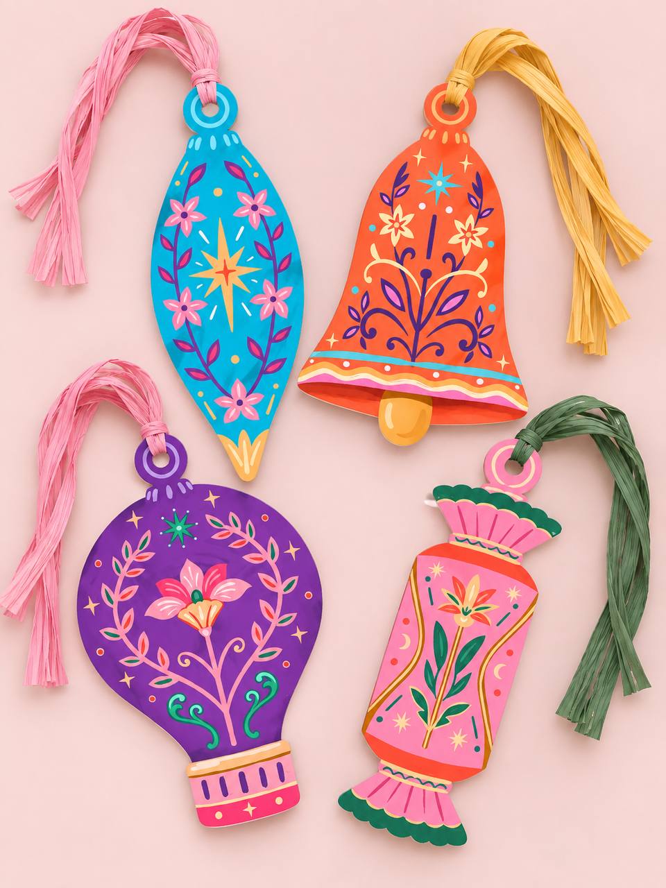 Four Talisman paper ornaments — Finial, Bell, Hot Air Balloon, and Candy — in blue, orange, purple, and pink folk-art floral designs, each with a colorful raffia tassel, on a pink background.