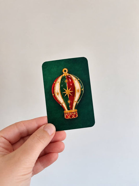 Hand holding a mini green Christmas card with an embossed red, green, and gold jeweled hot air balloon ornament illustration.