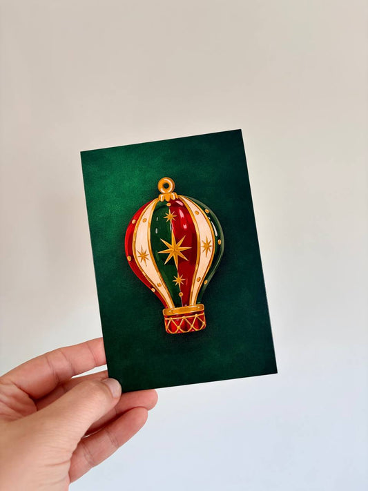 Hand holding a green Christmas greeting card with an embossed red, green, and gold jeweled hot air balloon ornament illustration.
