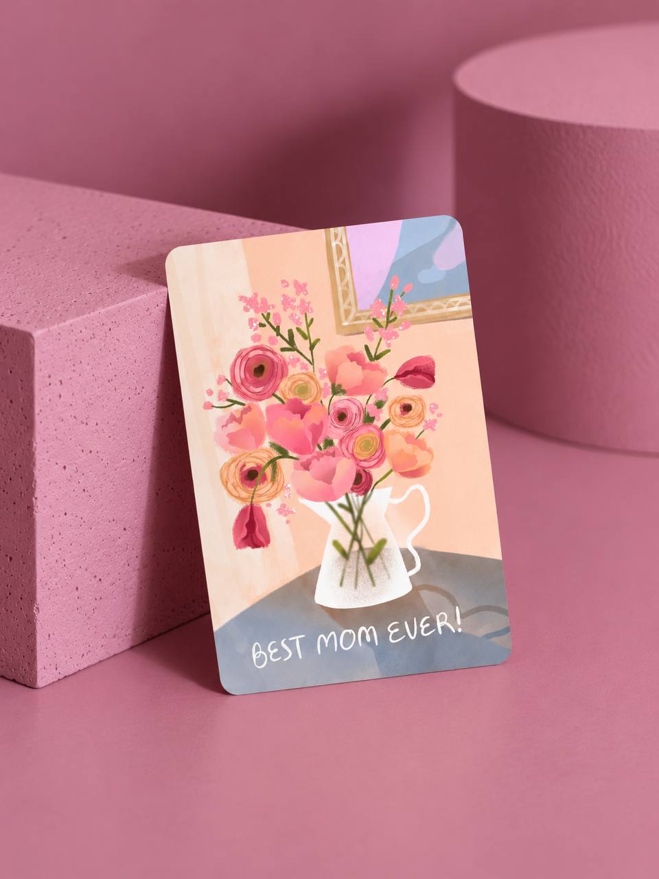 Greeting card illustrated with a pink and peach floral bouquet in a white pitcher and "Best Mom Ever!" text, propped against pink display blocks.
